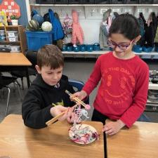 Two students learning to use chopsticks to pickup noodles.